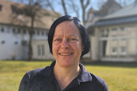 Eine Therapeutin der KIH im Freien auf einer Wiese mit einem Gebäude im Hintergrund und trägt dunkle Therapie-Arbeitskleidung.