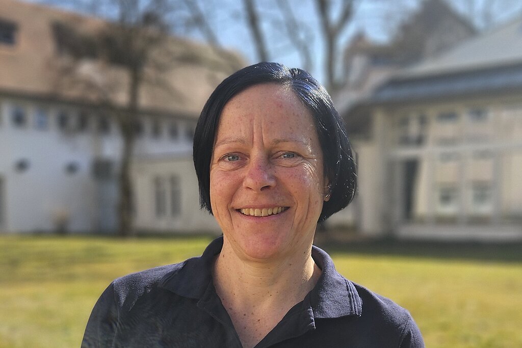 Eine Therapeutin der KIH im Freien auf einer Wiese mit einem Gebäude im Hintergrund und trägt dunkle Therapie-Arbeitskleidung.