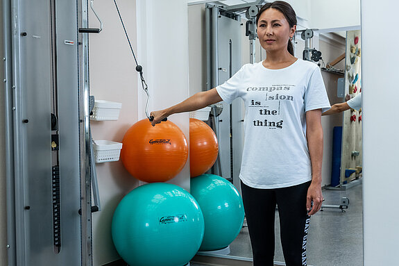 Eine Frau macht eine Übung in einem Trainingsraum mit medizinischen Trainingsgeräten und Gymnastikbällen.
