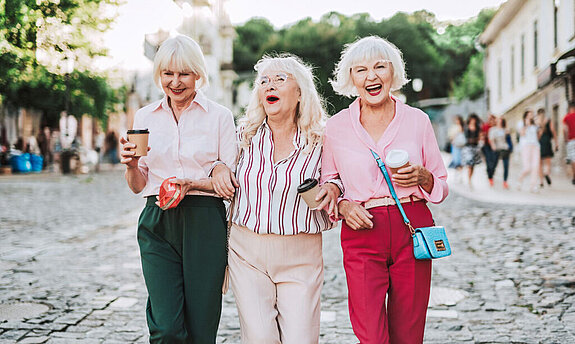 Drei Seniorinnen laufen lachend durch die Straße.