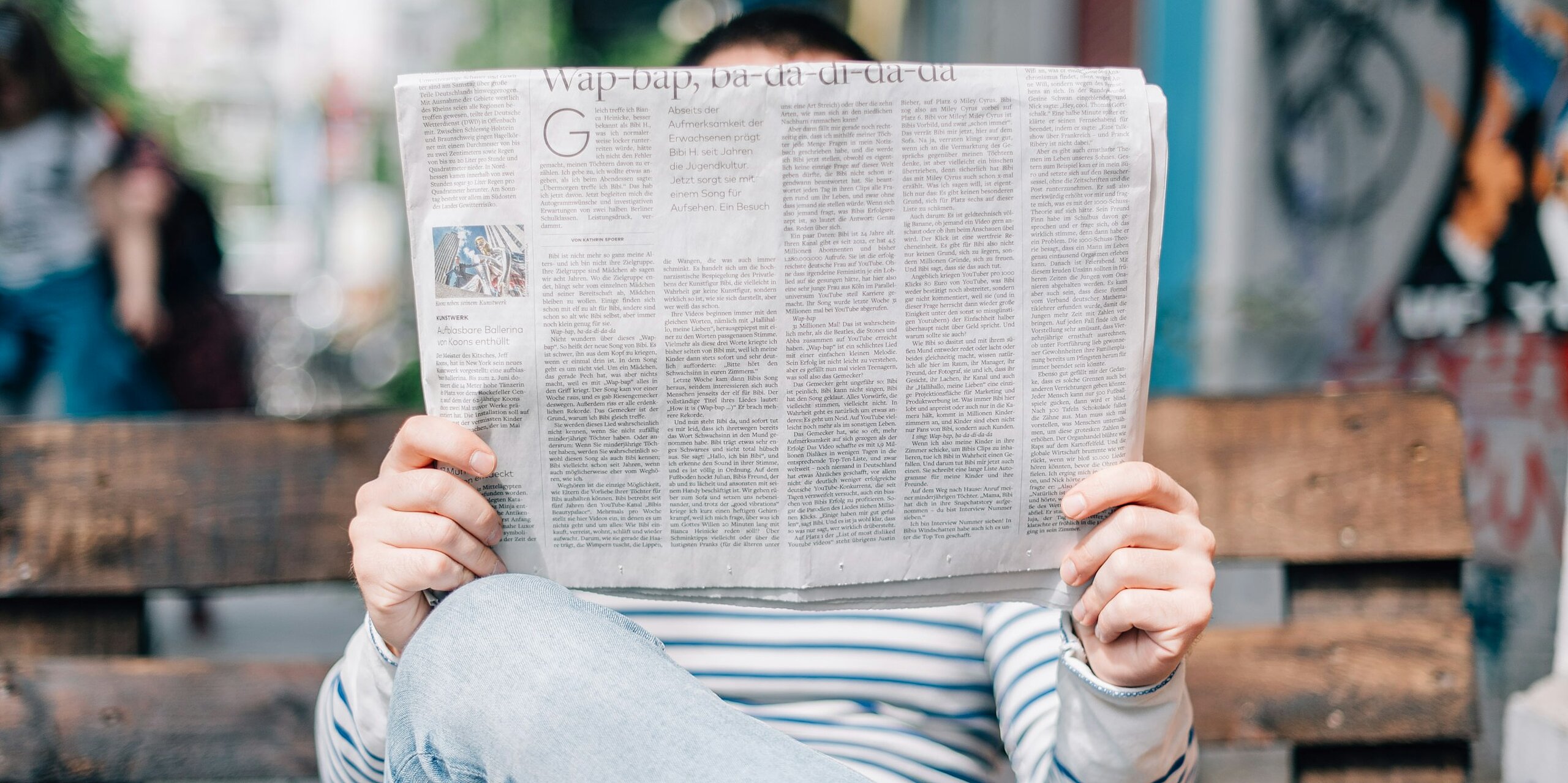 Ein Mensch ließt eine Zeitung auf einer Parkbank. Das Gesicht ist bedeckt von der Zeitung.