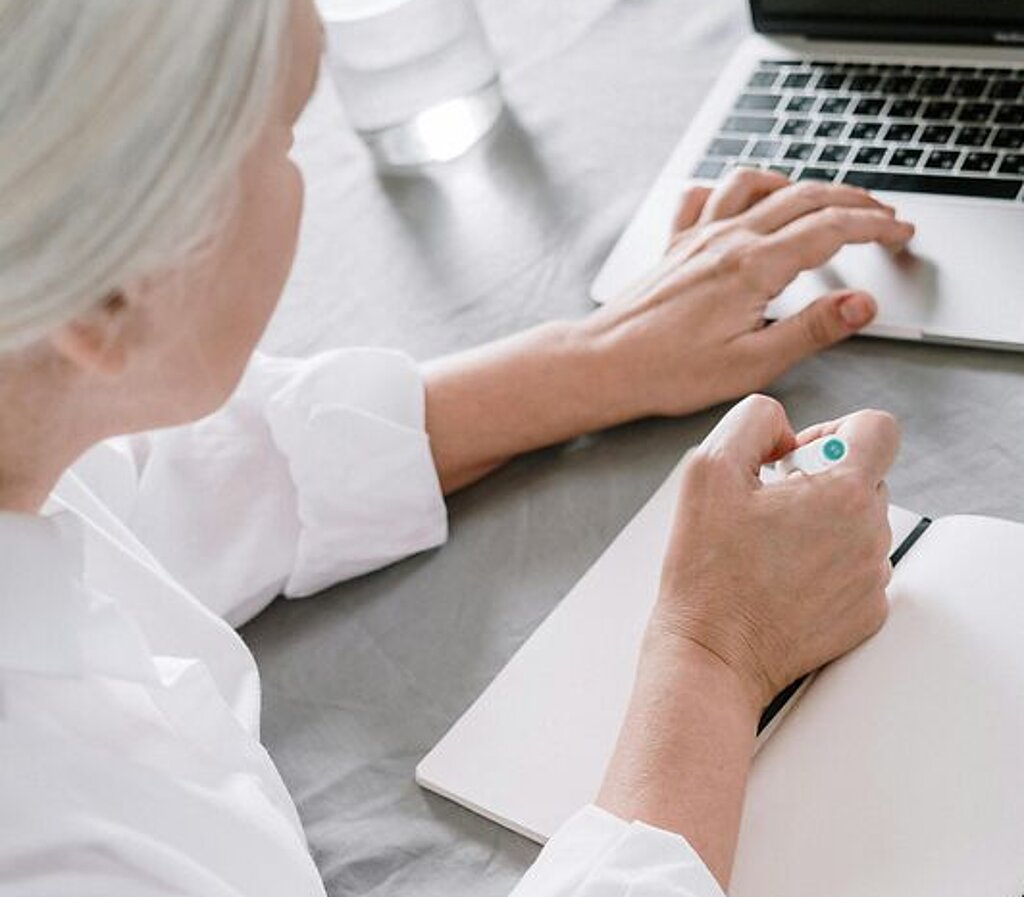 Frau am Laptop Eine ältere Frau sucht etwas am Laptop und macht sich dabei Notizen auf einen Block.