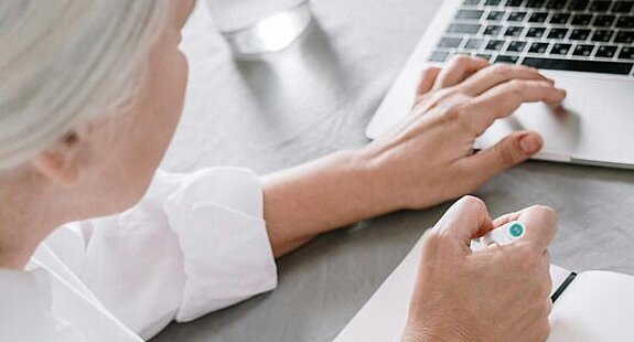 Frau recherchiert am Laptop. Eine ältere Frau sucht etwas am Laptop und macht sich dabei Notizen auf einen Block.