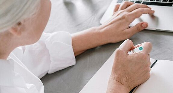 Frau am Laptop Eine ältere Frau sucht etwas am Laptop und macht sich dabei Notizen auf einen Block.