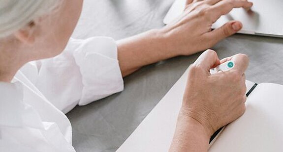 Frau am Laptop Eine ältere Frau sucht etwas am Laptop und macht sich dabei Notizen auf einen Block.