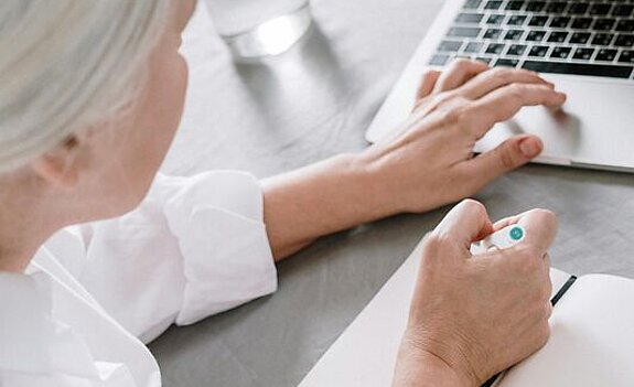 Frau am Laptop Eine ältere Frau sucht etwas am Laptop und macht sich dabei Notizen auf einen Block.