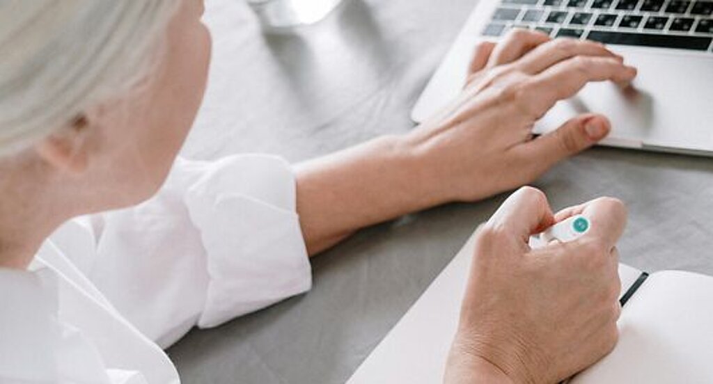 Frau am Laptop Eine ältere Frau sucht etwas am Laptop und macht sich dabei Notizen auf einen Block.