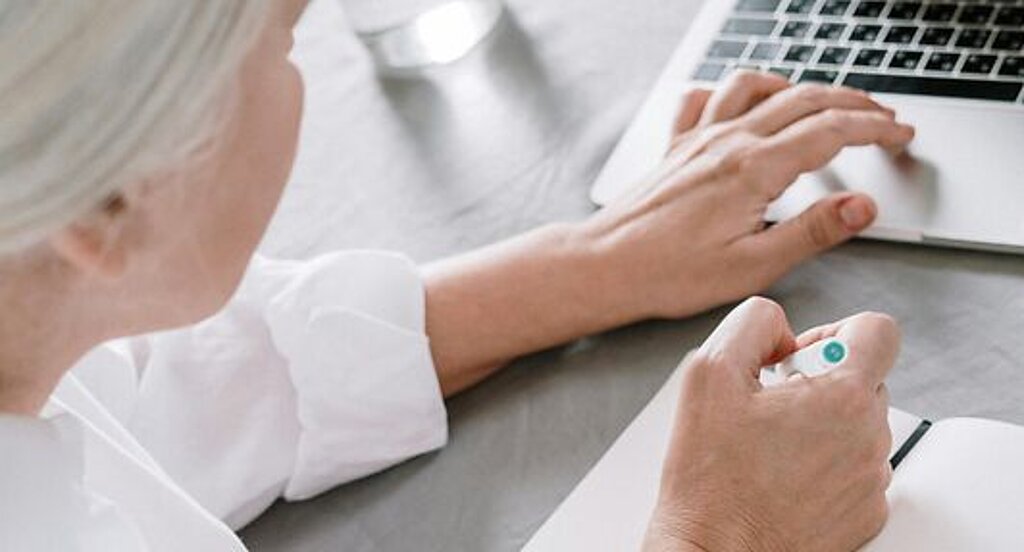 Frau am Laptop Eine ältere Frau sucht etwas am Laptop und macht sich dabei Notizen auf einen Block.