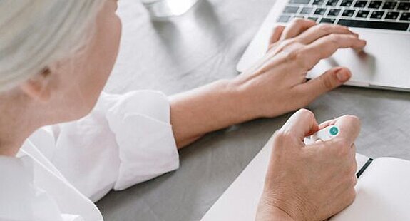 Frau am Laptop Eine ältere Frau sucht etwas am Laptop und macht sich dabei Notizen auf einen Block.