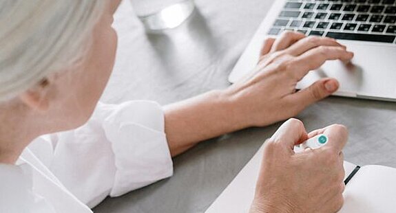 Frau recherchiert am Laptop. Eine ältere Frau sucht etwas am Laptop und macht sich dabei Notizen auf einen Block.