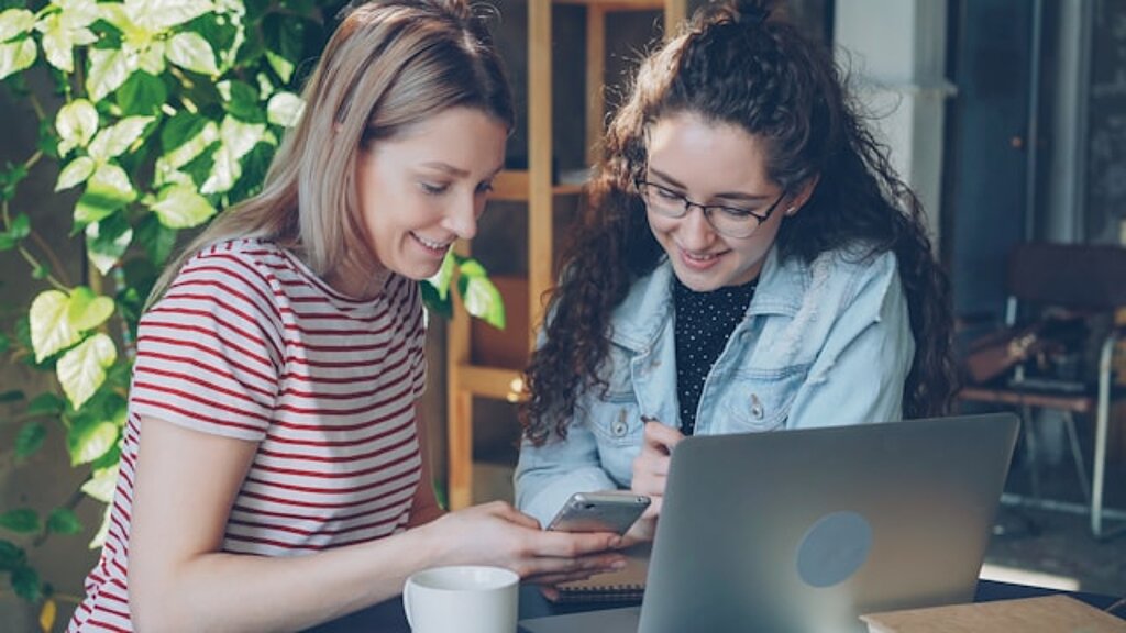 Zwei Studentinnen schauen gemeinsam in einen Laptop und ein Smartphone.