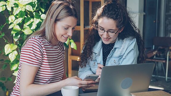 Zwei Studentinnen schauen gemeinsam in einen Laptop und ein Smartphone.