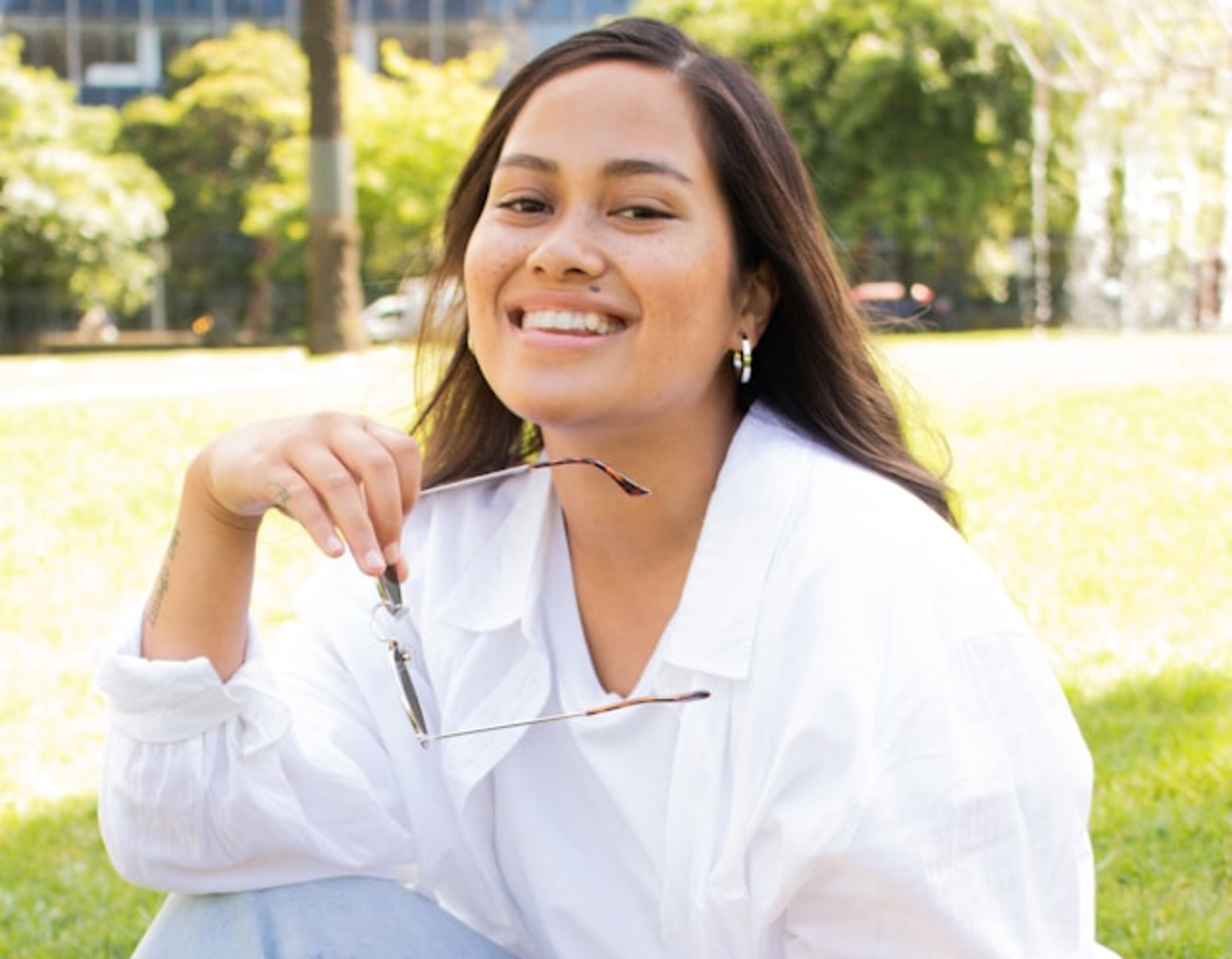Eine Studentin sitzt lachend im Park mit Tasche.