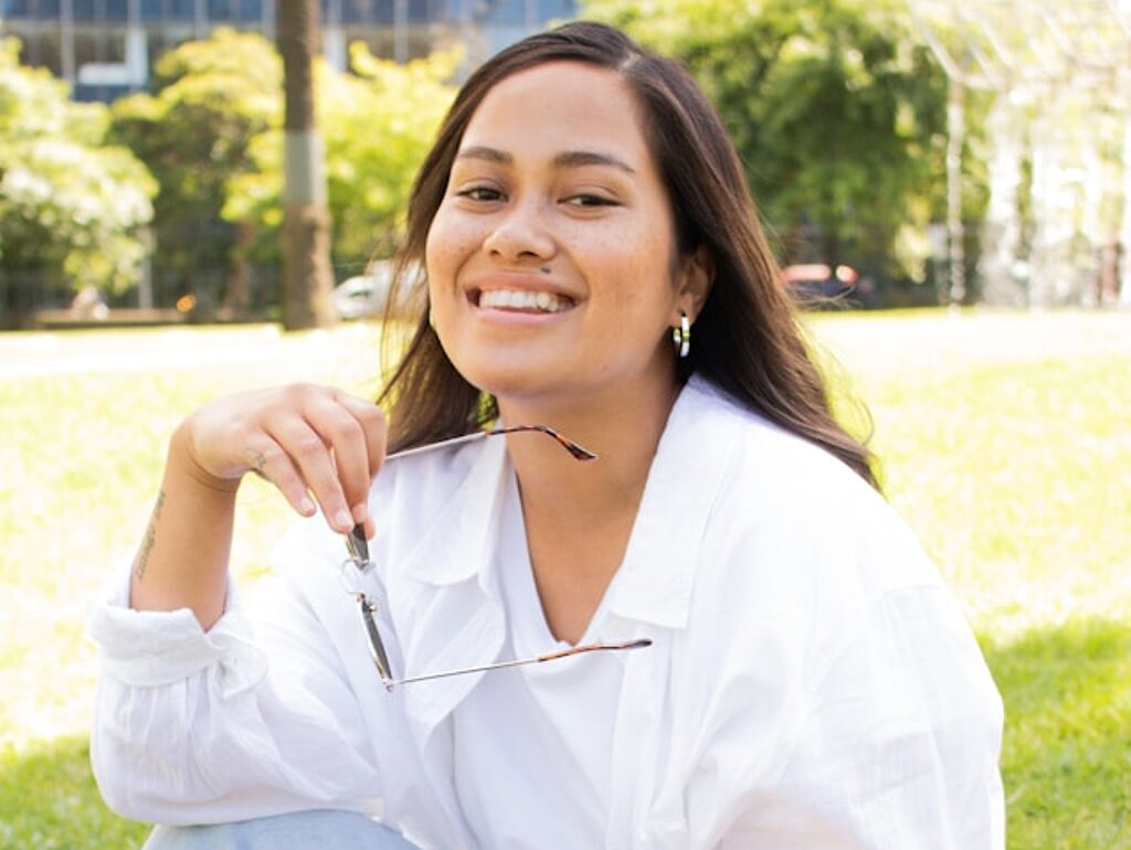 Eine Studentin sitzt lachend im Park mit Tasche.
