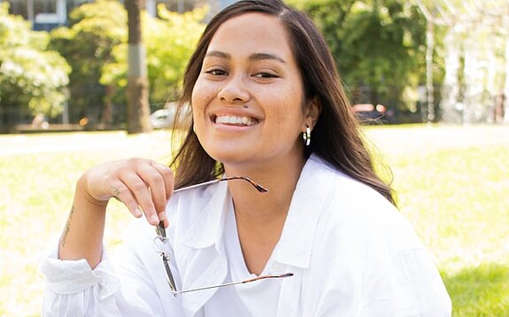 Eine Studentin sitzt lachend im Park mit Tasche.