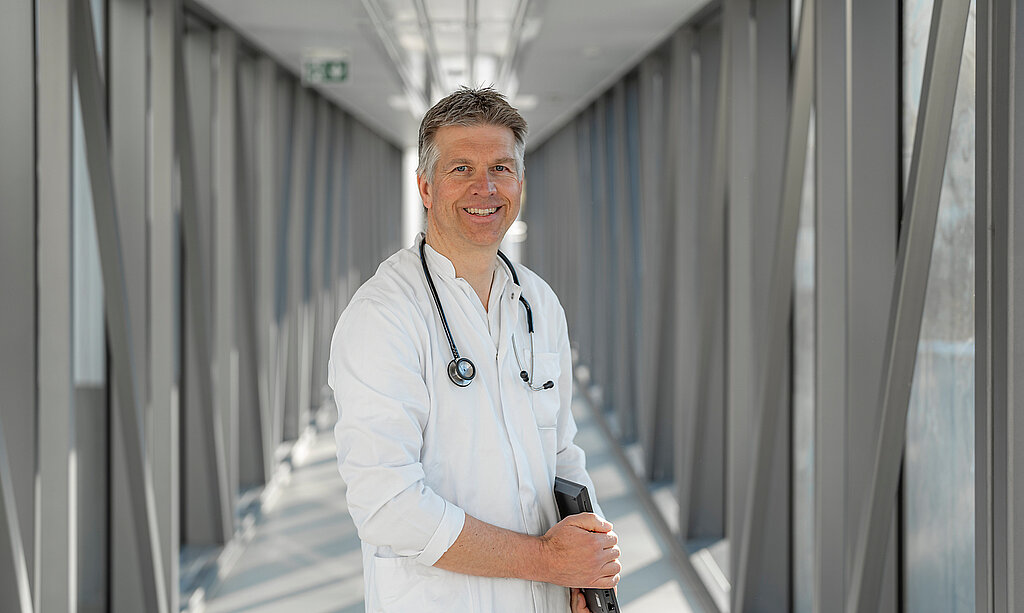 Ein lächelnder Arzt steht mit einer Mappe im Gang.