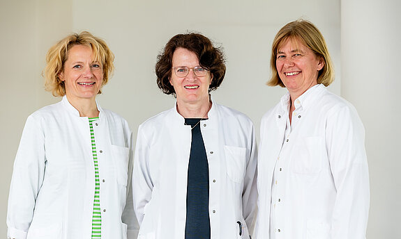 Drei Ärztinnen mit weißem Kittel lächeln in die Kamera.