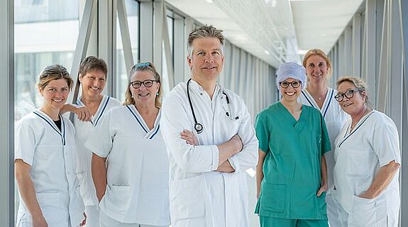 Arzt und ein Team aus Pflegekräften stehen gemeinsam in einem hellen Glasgang.