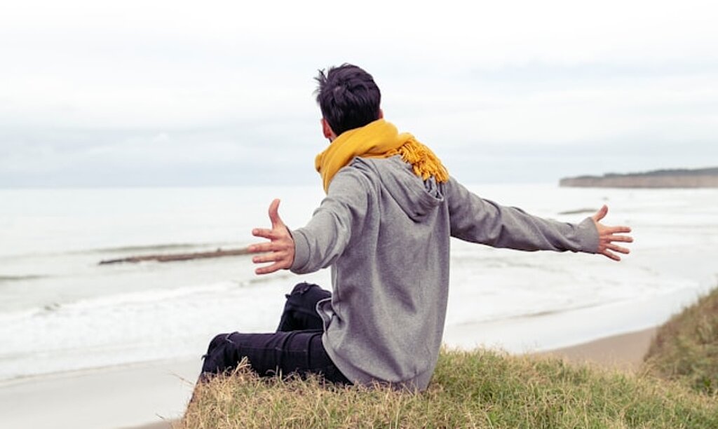 Ein Mann sitzt auf einem Hand und breitet die Arme aus, im Hintergrund sieht man das Meer.