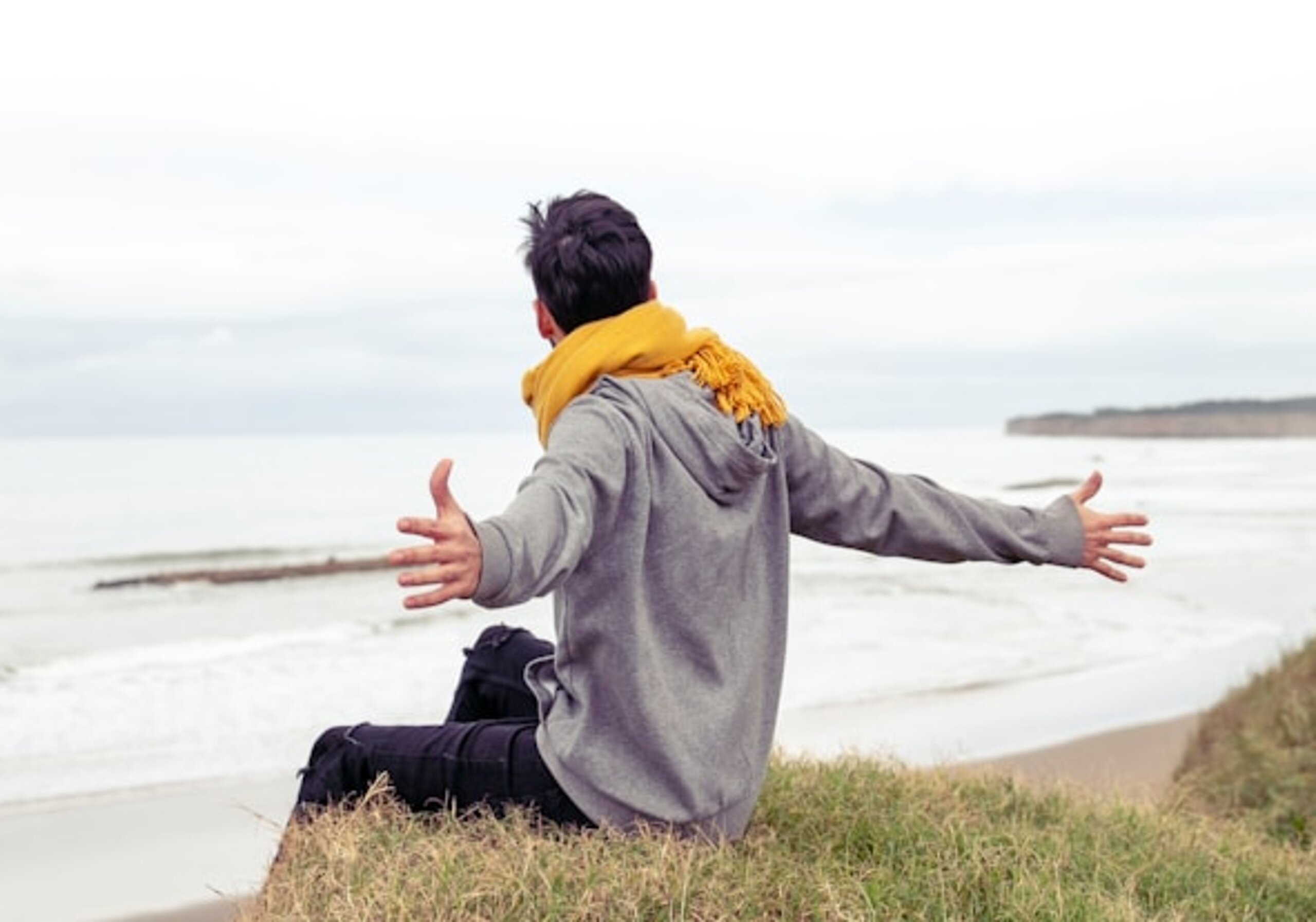 Ein Mann sitzt auf einem Hand und breitet die Arme aus, im Hintergrund sieht man das Meer.