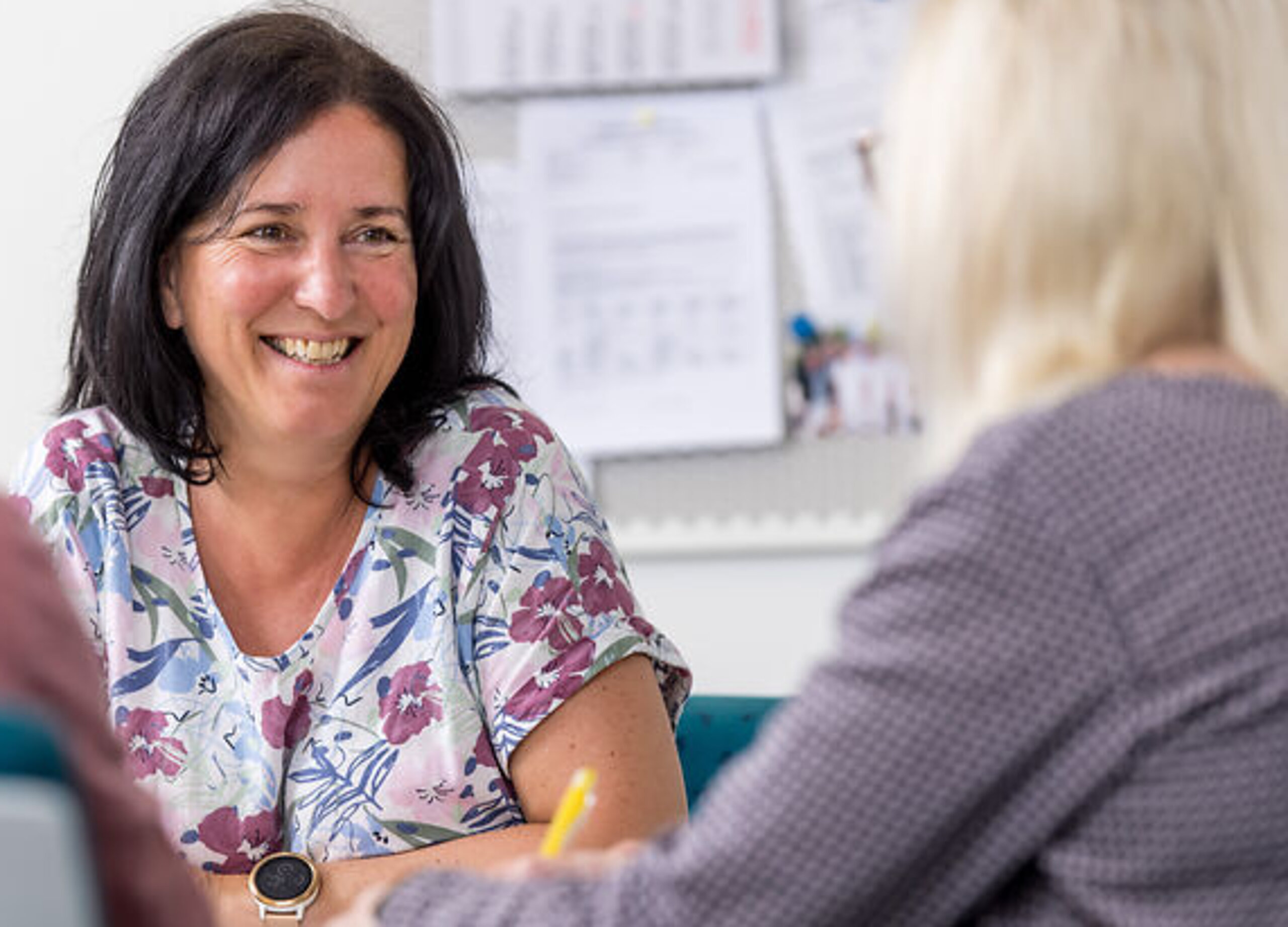 Eine Mitarbeiterin unterhält sich freundlich mit zwei Patienten am Schreibtisch.