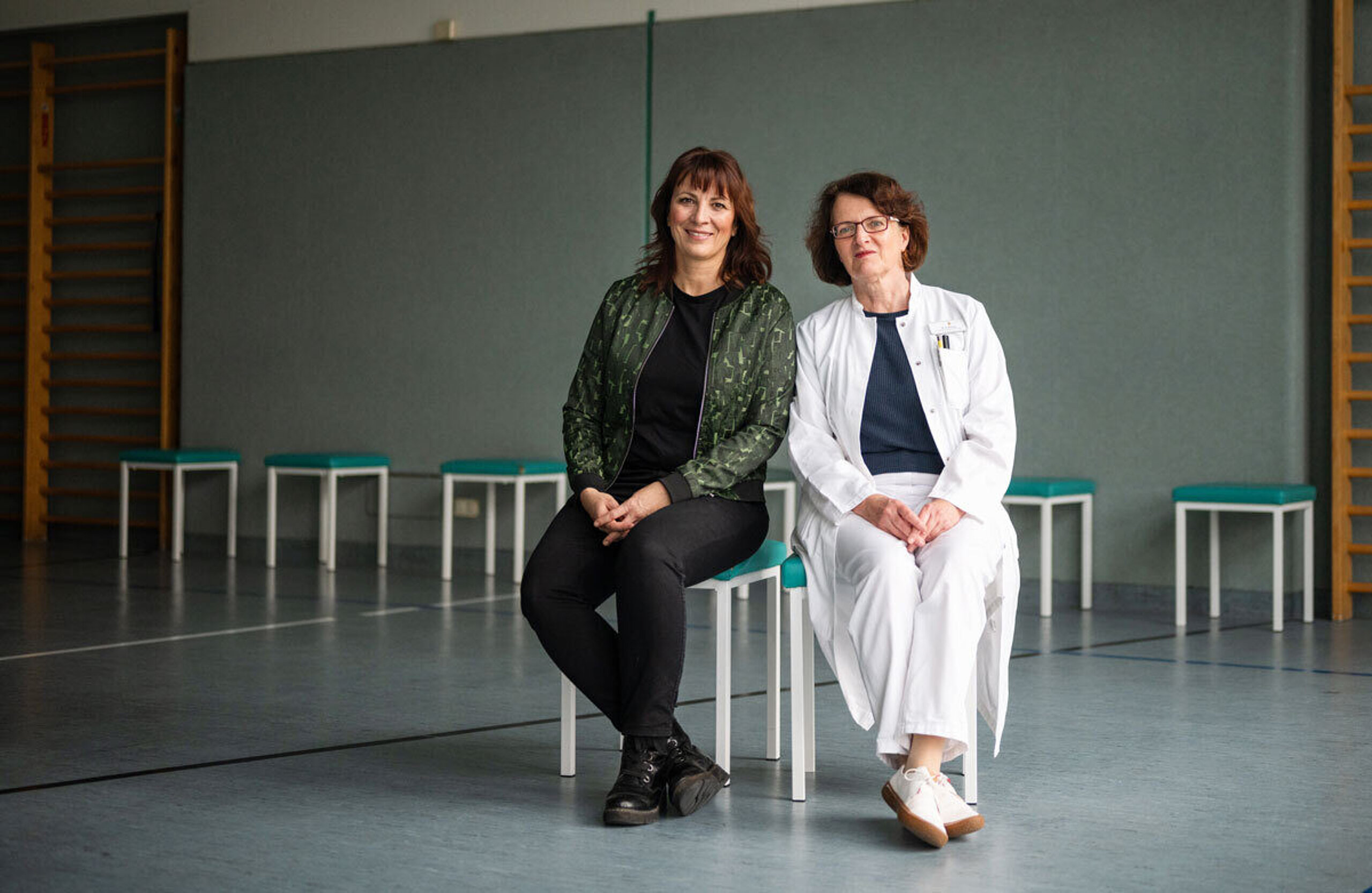 Heilsamer Sport Dr. Ivonne Rudolph (links) und Dr. Bettine Bilsing sitzen in einer Sporthalle auf Stühlen.
