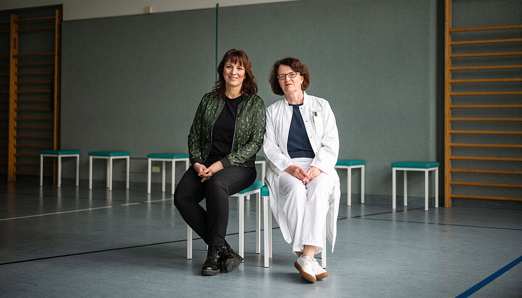 Heilsamer Sport Dr. Ivonne Rudolph (links) und Dr. Bettine Bilsing sitzen in einer Sporthalle auf Stühlen.