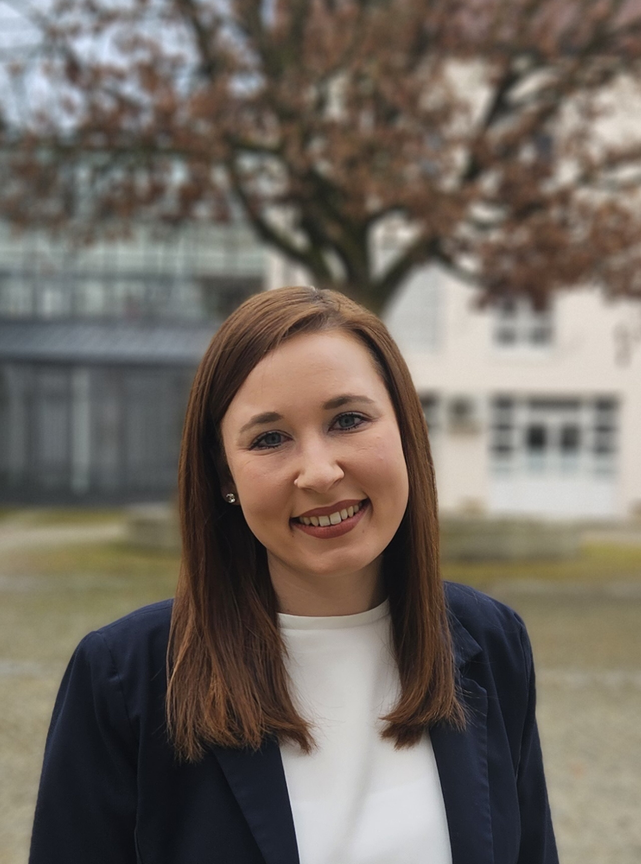 Die Personalreferentin der KIH mit weißer Bluse und dunklem Jackett steht draußen auf einem Platz mit Pflastersteinen und Gebäuden im Hintergrund.