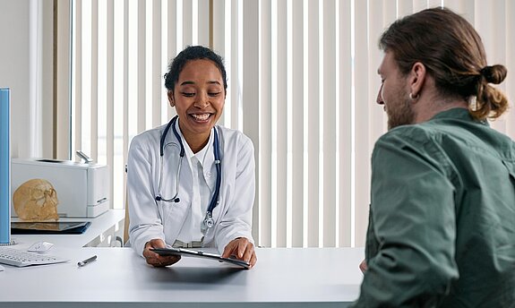 Eine Ärztin im Gespräch mit einem Patienten in einem modernen Praxisraum am Schreibtisch.