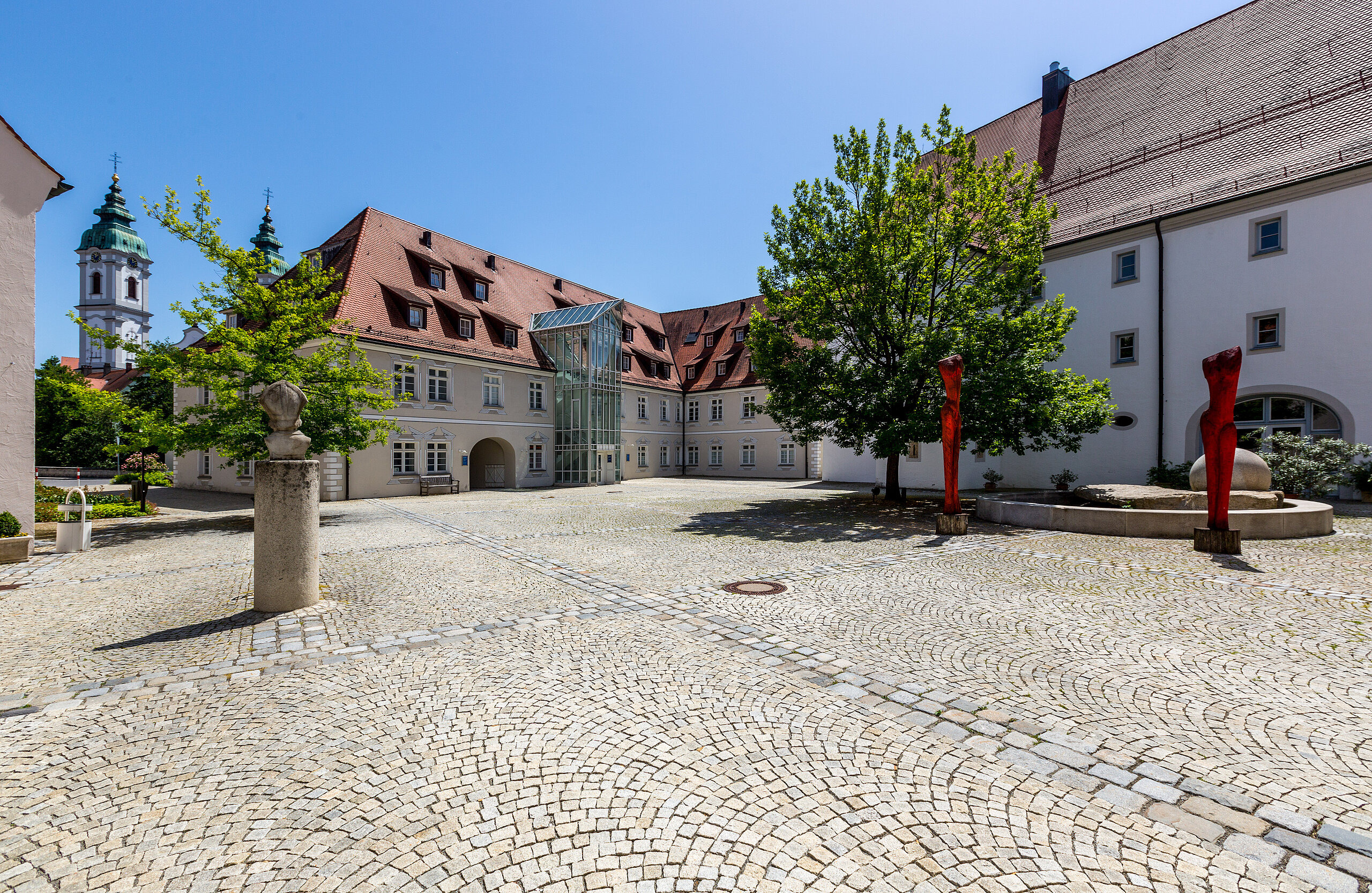 Gepflasterter Innenhof der Klinik im Hofgarten in Bad Waldsee, umgeben von historischen Gebäuden mit roten Dächern, Bäumen und blauem Himmel im Hintergrund