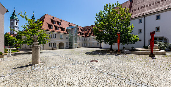 Gepflasterter Innenhof der Klinik im Hofgarten in Bad Waldsee, umgeben von historischen Gebäuden mit roten Dächern, Bäumen und blauem Himmel im Hintergrund