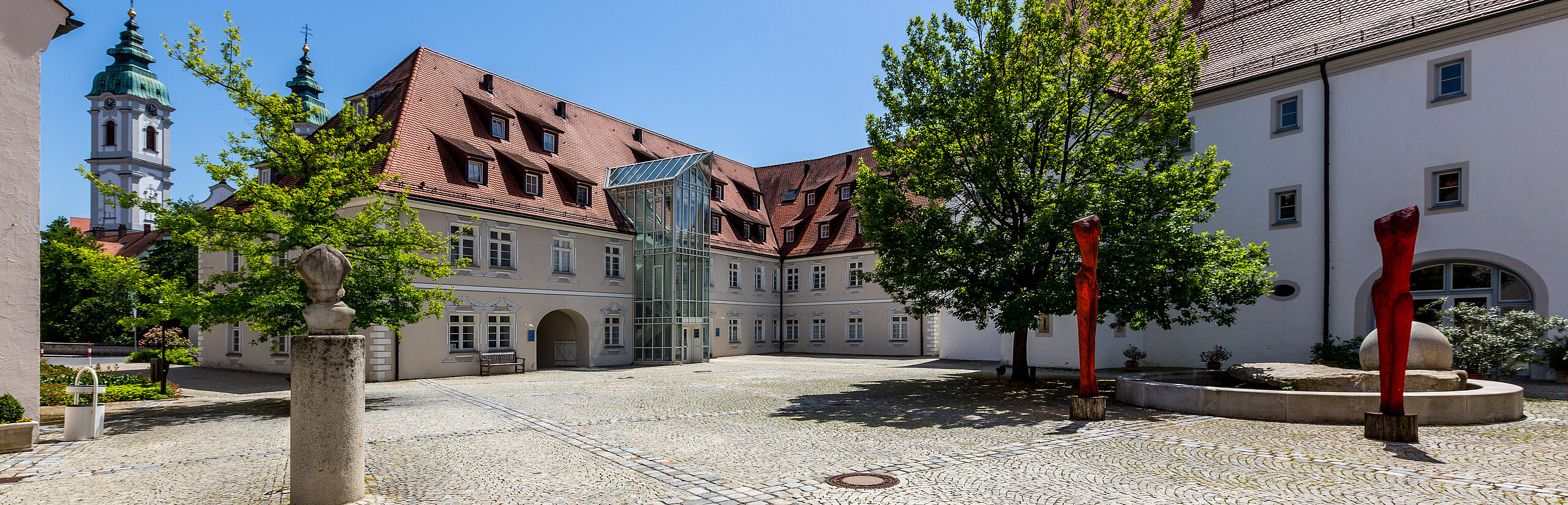Gepflasterter Innenhof der Klinik im Hofgarten in Bad Waldsee, umgeben von historischen Gebäuden mit roten Dächern, Bäumen und blauem Himmel im Hintergrund