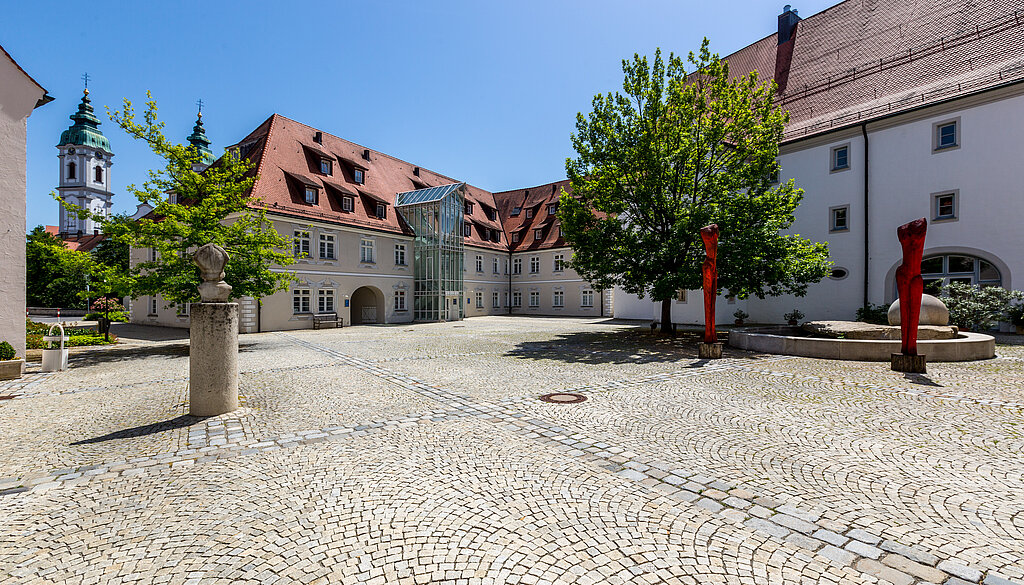 Gepflasterter Innenhof der Klinik im Hofgarten in Bad Waldsee, umgeben von historischen Gebäuden mit roten Dächern, Bäumen und blauem Himmel im Hintergrund