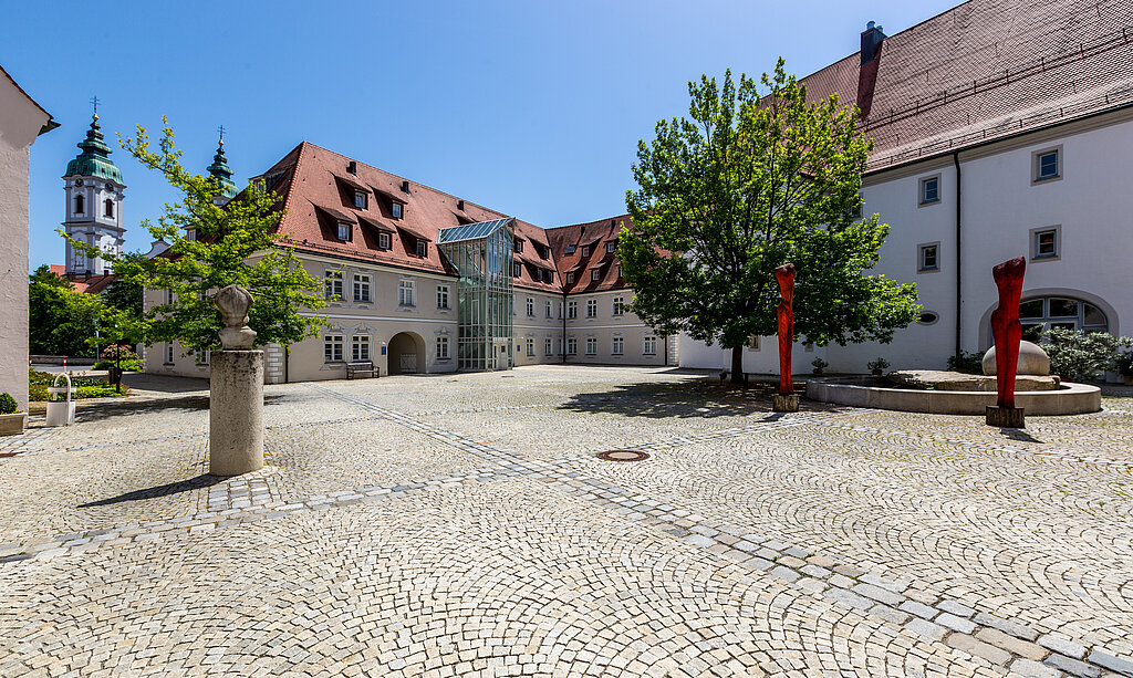 Gepflasterter Innenhof der Klinik im Hofgarten in Bad Waldsee, umgeben von historischen Gebäuden mit roten Dächern, Bäumen und blauem Himmel im Hintergrund