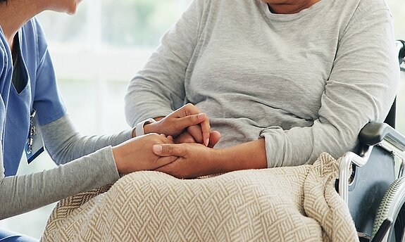Eine Pflegerin hält vertrauensvoll die Hand einer Patientin im Rollstuhl.