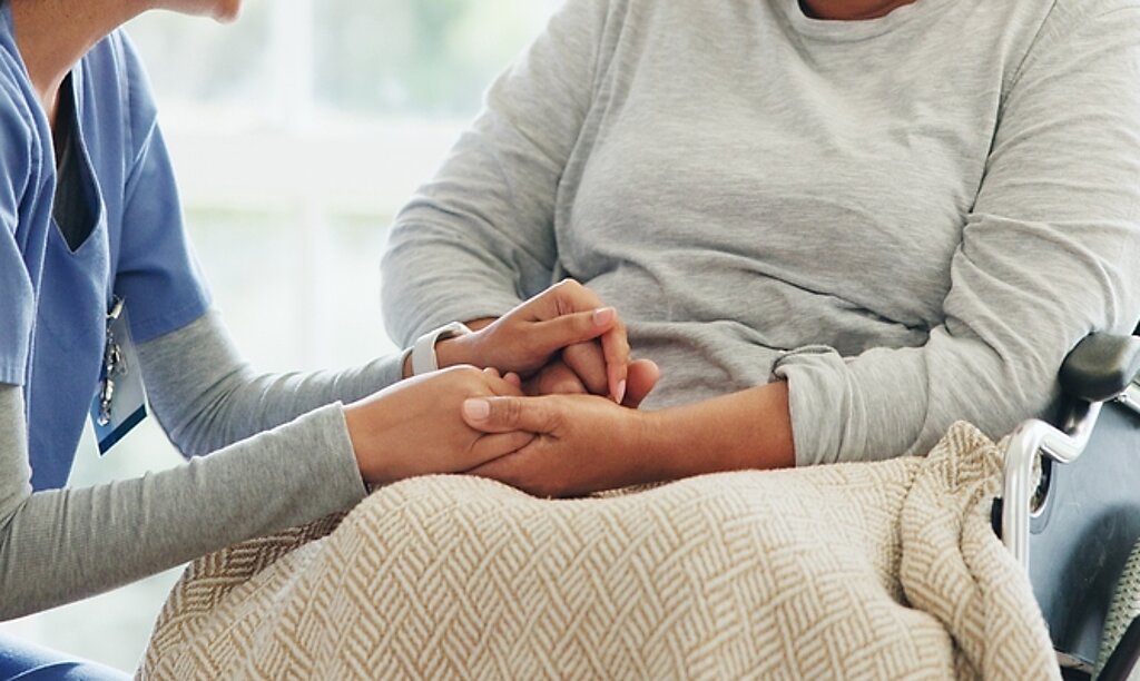 Eine Pflegerin hält vertrauensvoll die Hand einer Patientin im Rollstuhl.