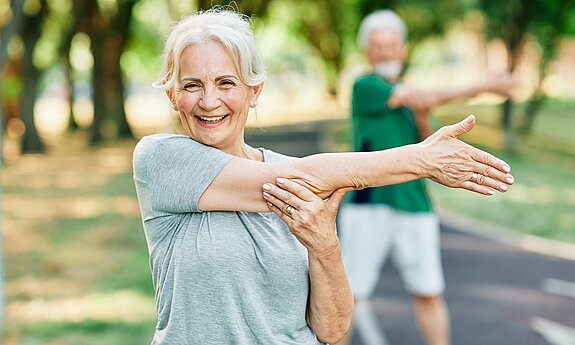 Ältere Frau macht im Park eine Dehnübung mit ausgestrecktem Arm und lächelt in die Kamera