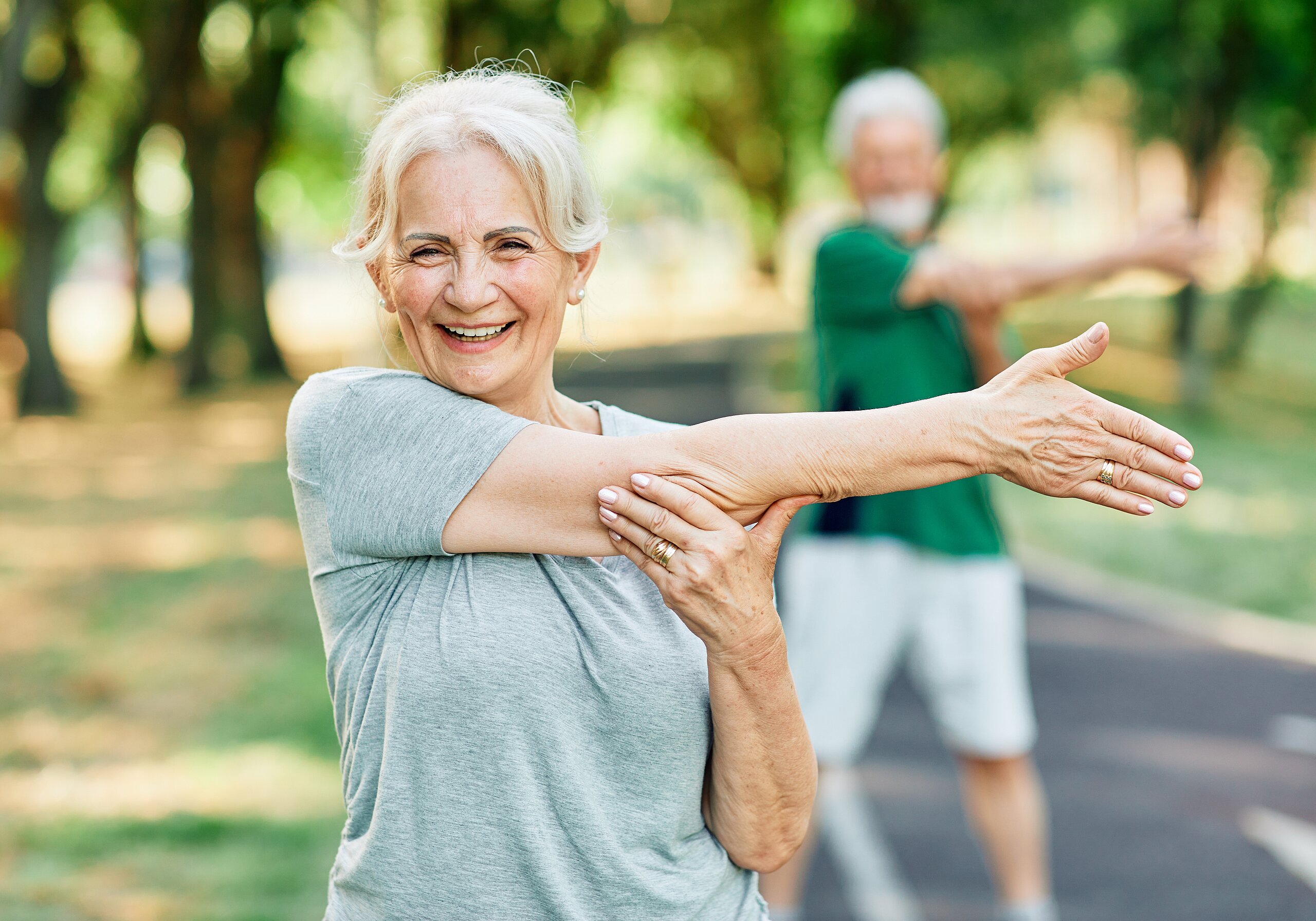 Ältere Frau macht im Park eine Dehnübung mit ausgestrecktem Arm und lächelt in die Kamera
