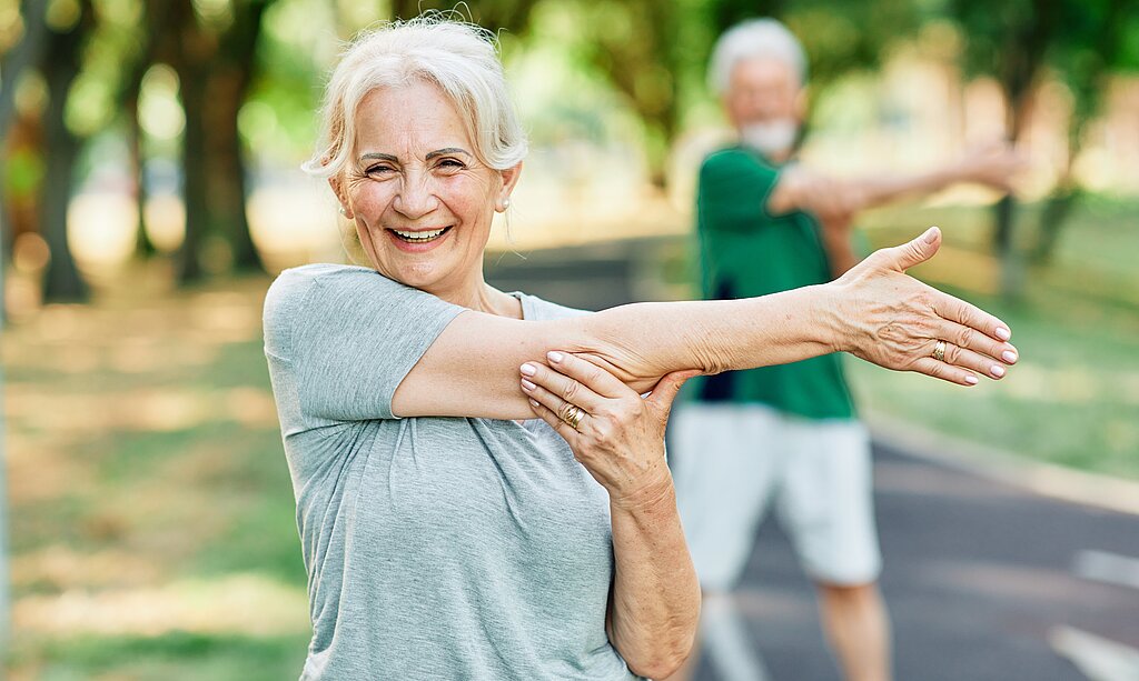Ältere Frau macht im Park eine Dehnübung mit ausgestrecktem Arm und lächelt in die Kamera