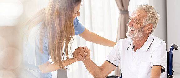 Ein älterer Patient im Rollstuhl wird freundlich von einer Pflegekraft angesprochen und betreut.