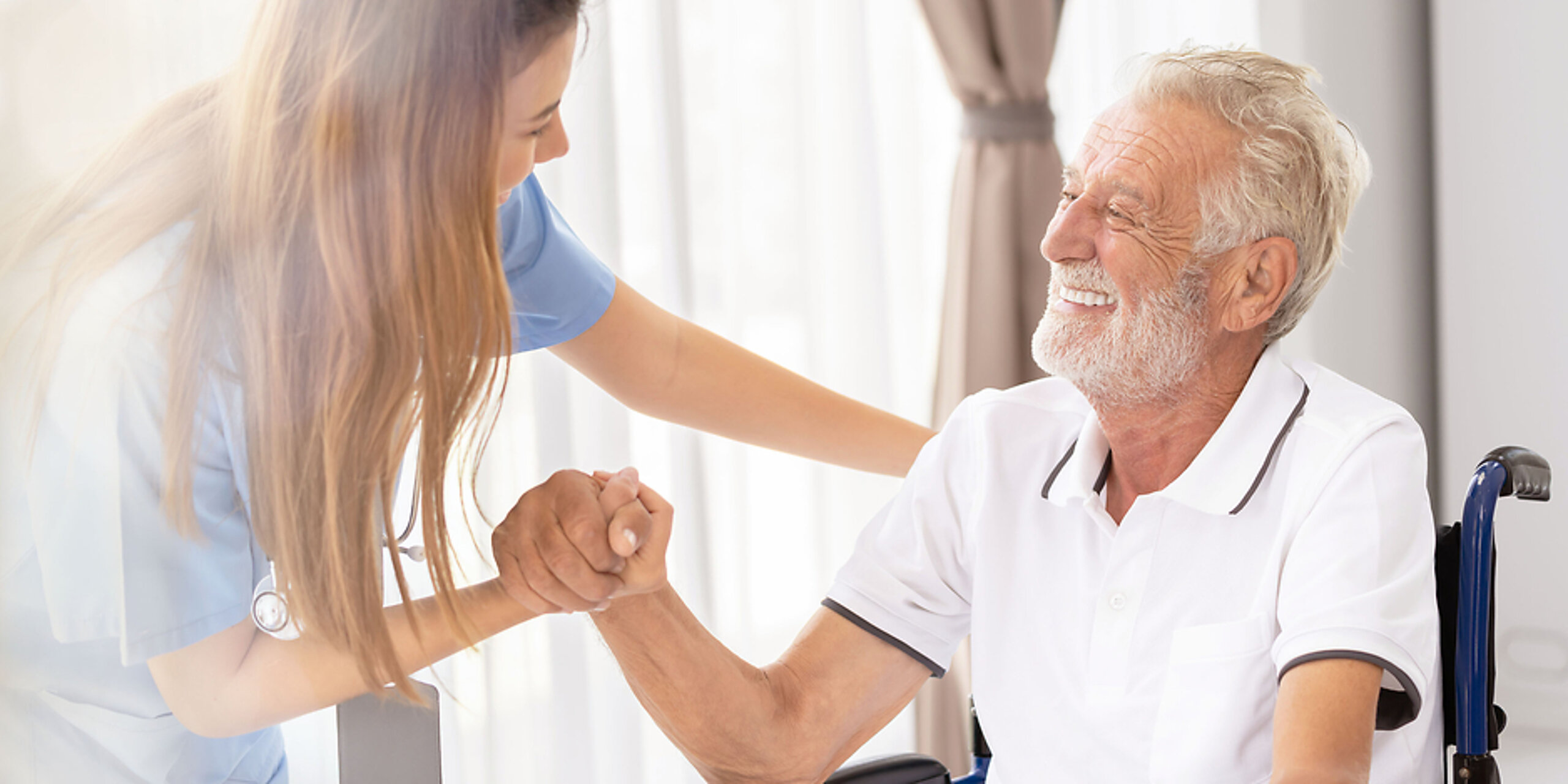 Ein älterer Patient im Rollstuhl wird freundlich von einer Pflegekraft angesprochen und betreut.