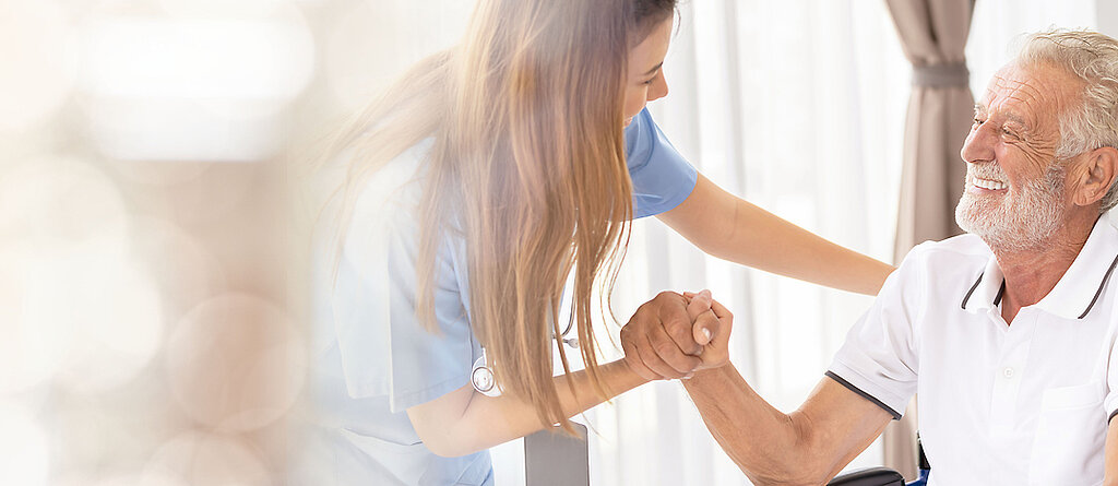 Ein älterer Patient im Rollstuhl wird freundlich von einer Pflegekraft angesprochen und betreut.