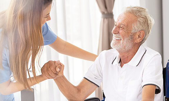 Ein älterer Patient im Rollstuhl wird freundlich von einer Pflegekraft angesprochen und betreut.