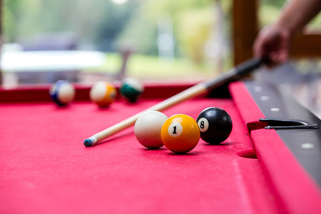 Das Bild zeigt einen Ausschnitt eines Billard-Tisches mit Kugeln, eine Hand hält einen Billardqueue.
