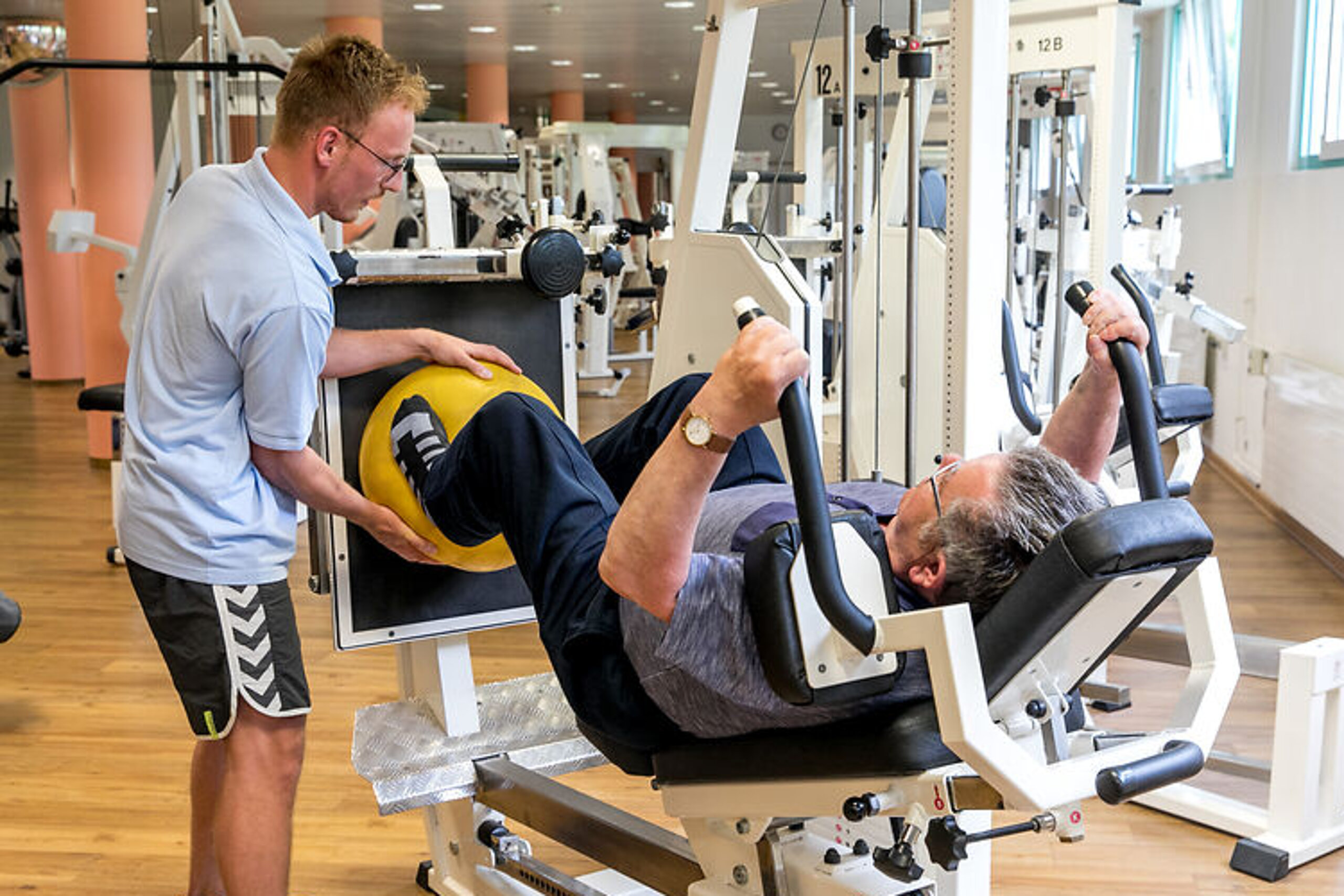 Ein Therapeut unterstützt einen Rehabilitanden beim Training an einer Beinpresse.