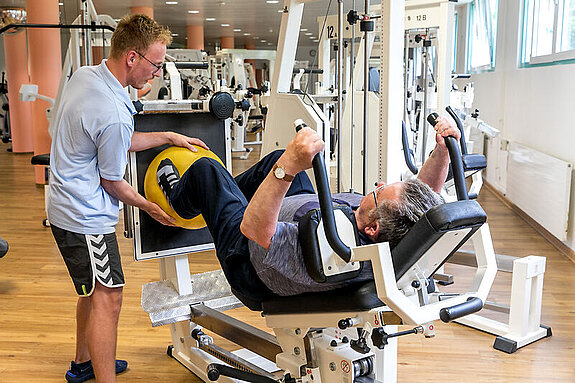 Ein Therapeut unterstützt einen Rehabilitanden beim Training an einer Beinpresse.
